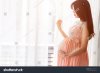 pregnant-woman-in-pink-dress-standing-close-up-a-window-in-bedroom-762919777.jpg